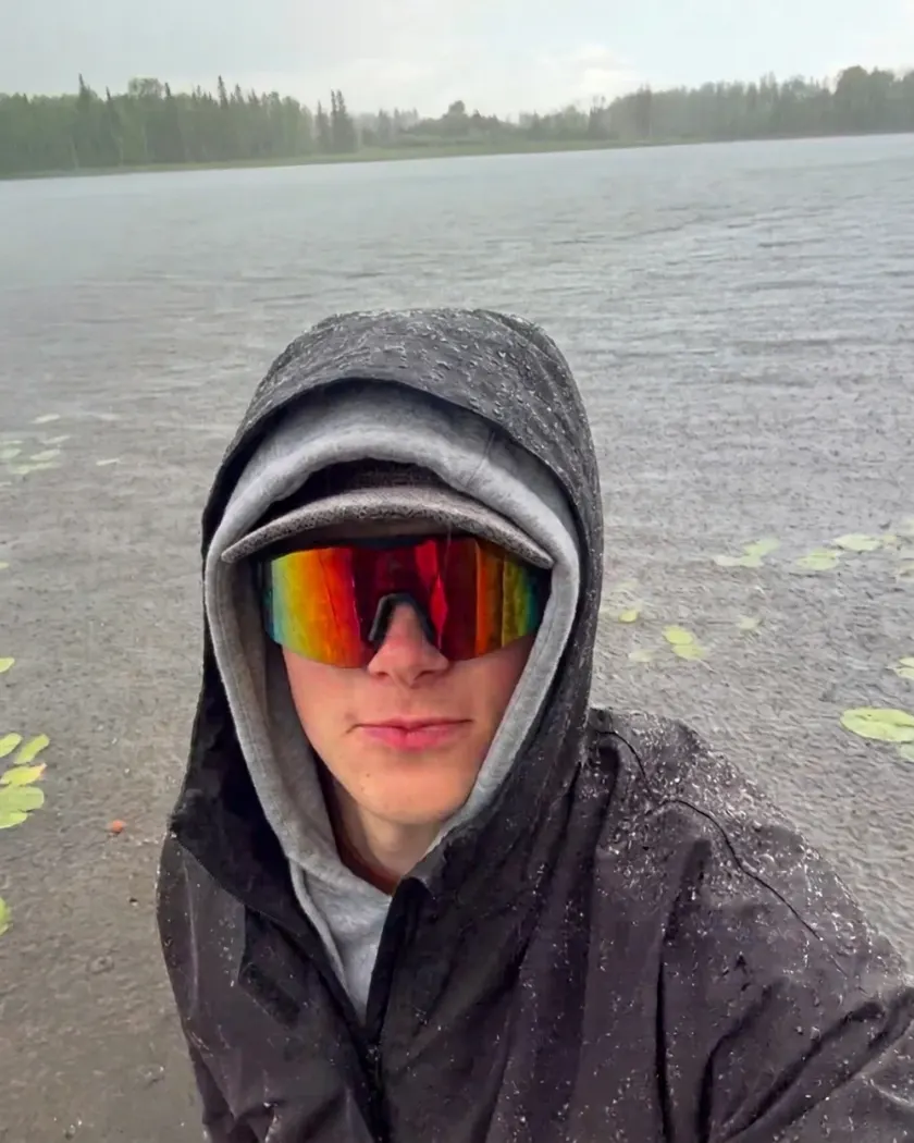 Petras Liubertas by a lake in the pouring rain, hood up, in mirrored sports sunglasses